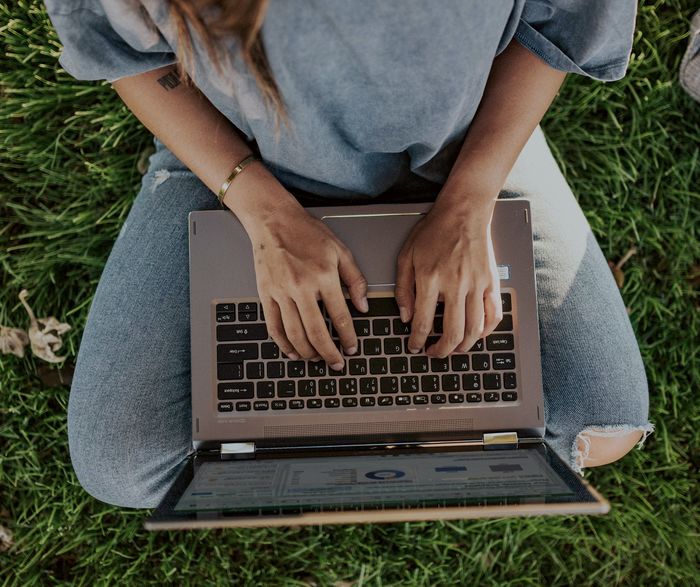 Eine Person sitzt im Gras. Sie hat einen Laptop auf ihrem Schoß und schreibt.